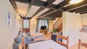 A spacious living room with a high ceiling, exposed beams, a dining area with a table and chairs, a television, and a staircase leading to another level. - Host & Stay | Providence Cottage (Woolacombe)