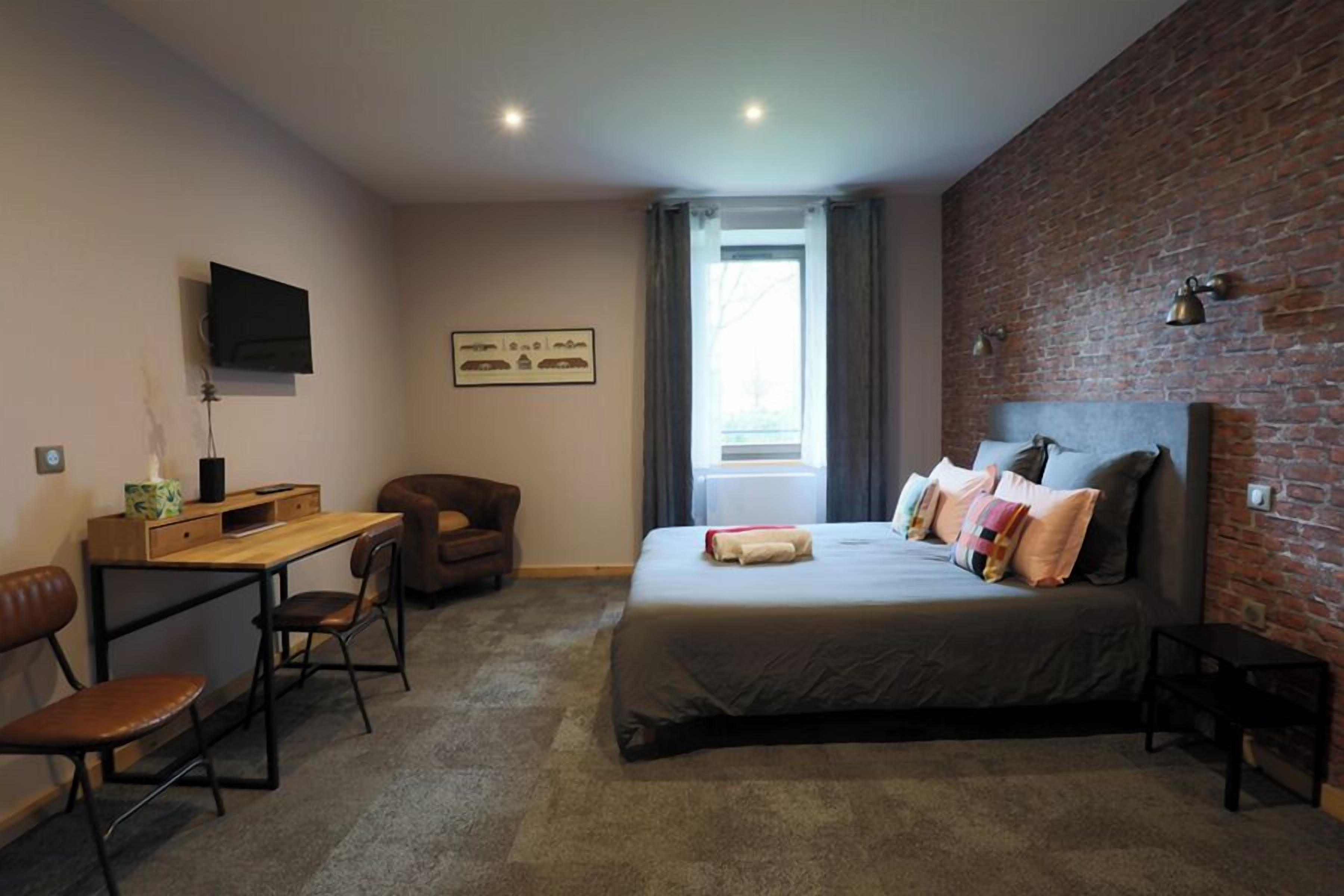 A bedroom with a bed, a desk, a chair, a TV, and a brick wall.