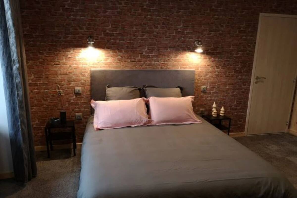 Une chambre avec un mur en briques, un lit avec deux oreillers, des tables de chevet et une porte.
