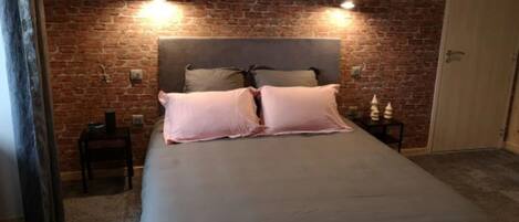 A bedroom with a brick wall, a bed with two pillows, bedside tables, and a door.
