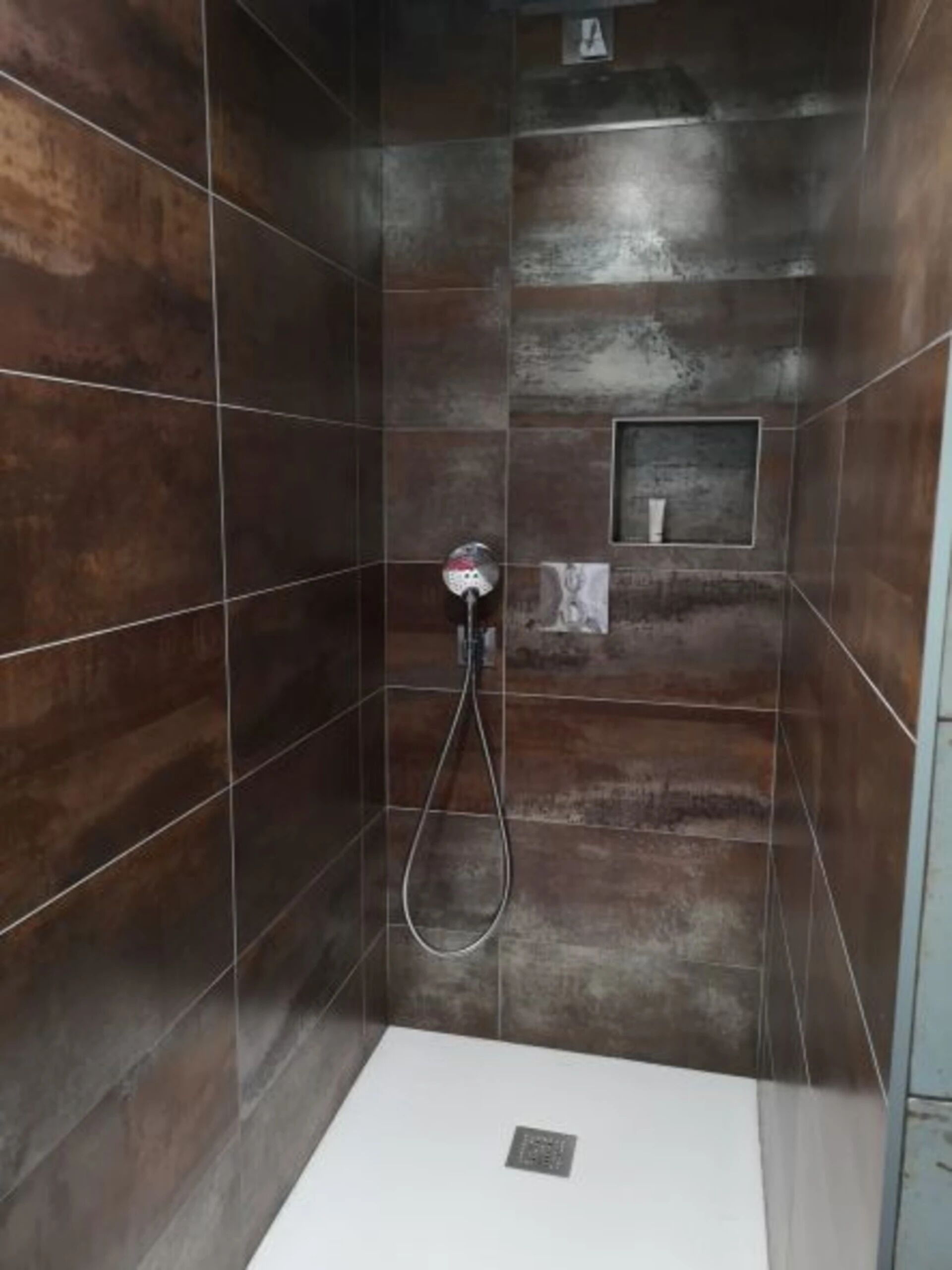 A shower stall with dark brown tiles, a handheld showerhead, and a small shelf with toiletries.