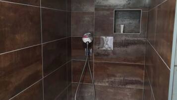 A shower stall with dark brown tiles, a handheld showerhead, and a small shelf with toiletries.