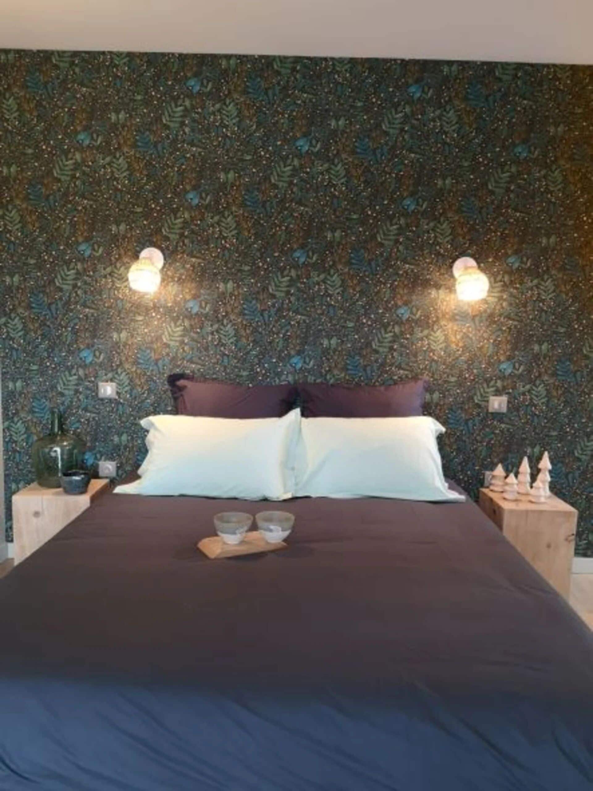 Ein Schlafzimmer mit gemustertem Wandpapier, einem Bett mit zwei Kissen, Nachttischen mit Dekoration und Wandleuchten.
