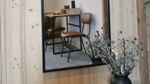 Een kamer met een grote spiegel, een houten wand, een bureau met stoel en een vaas met gedroogde bloemen.