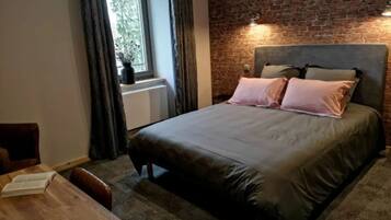 Un dormitorio con pared de ladrillo, una cama con dos almohadas, una ventana con cortinas y una mesa de madera con un libro.