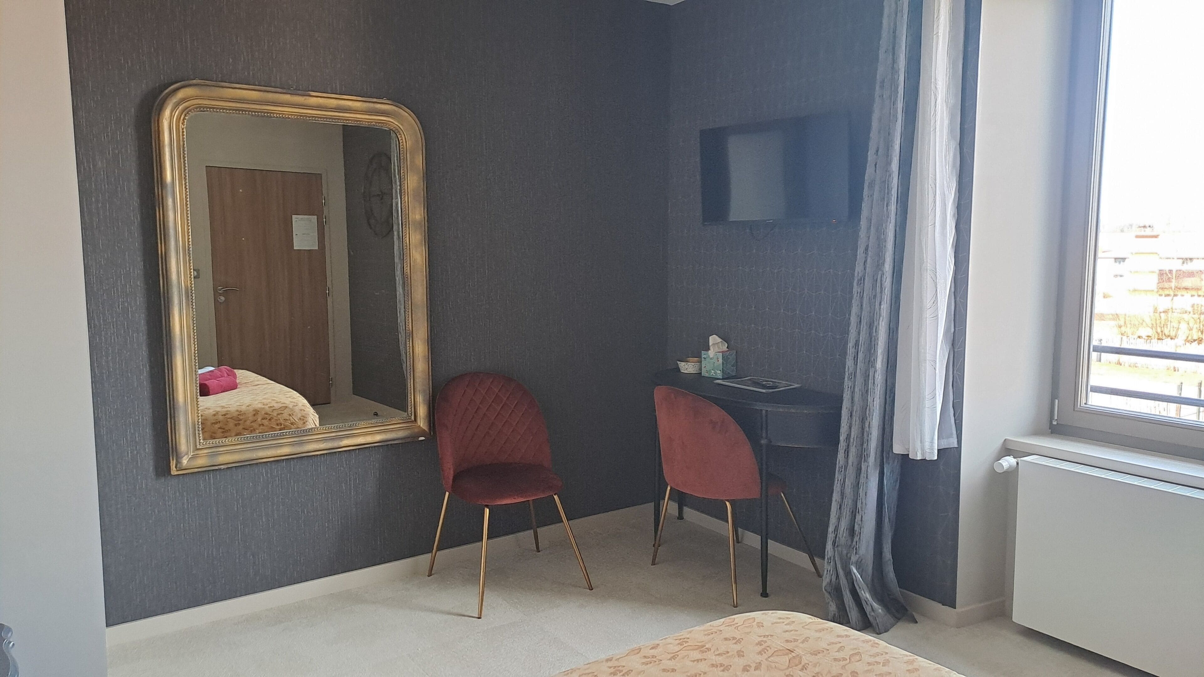 A room with a large mirror, two red chairs, a small table with a computer, and a window with curtains.
