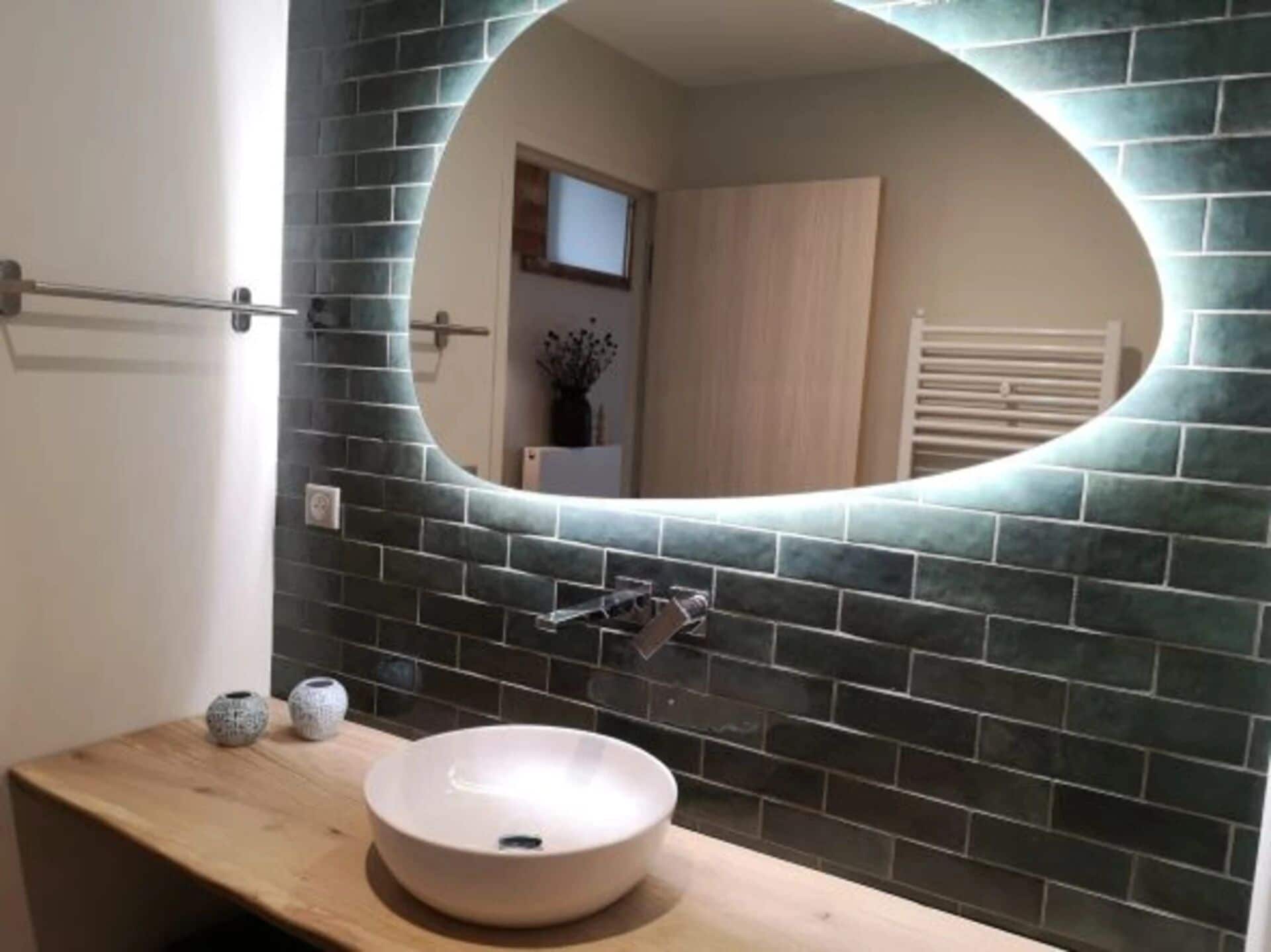 Une salle de bain avec un grand miroir rond, un pare-feu en carreaux de couleur sombre et un meuble-lavabo en bois avec un lavabo blanc.