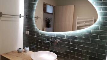 Une salle de bain avec un grand miroir rond, un pare-feu en carreaux de couleur sombre et un meuble-lavabo en bois avec un lavabo blanc.