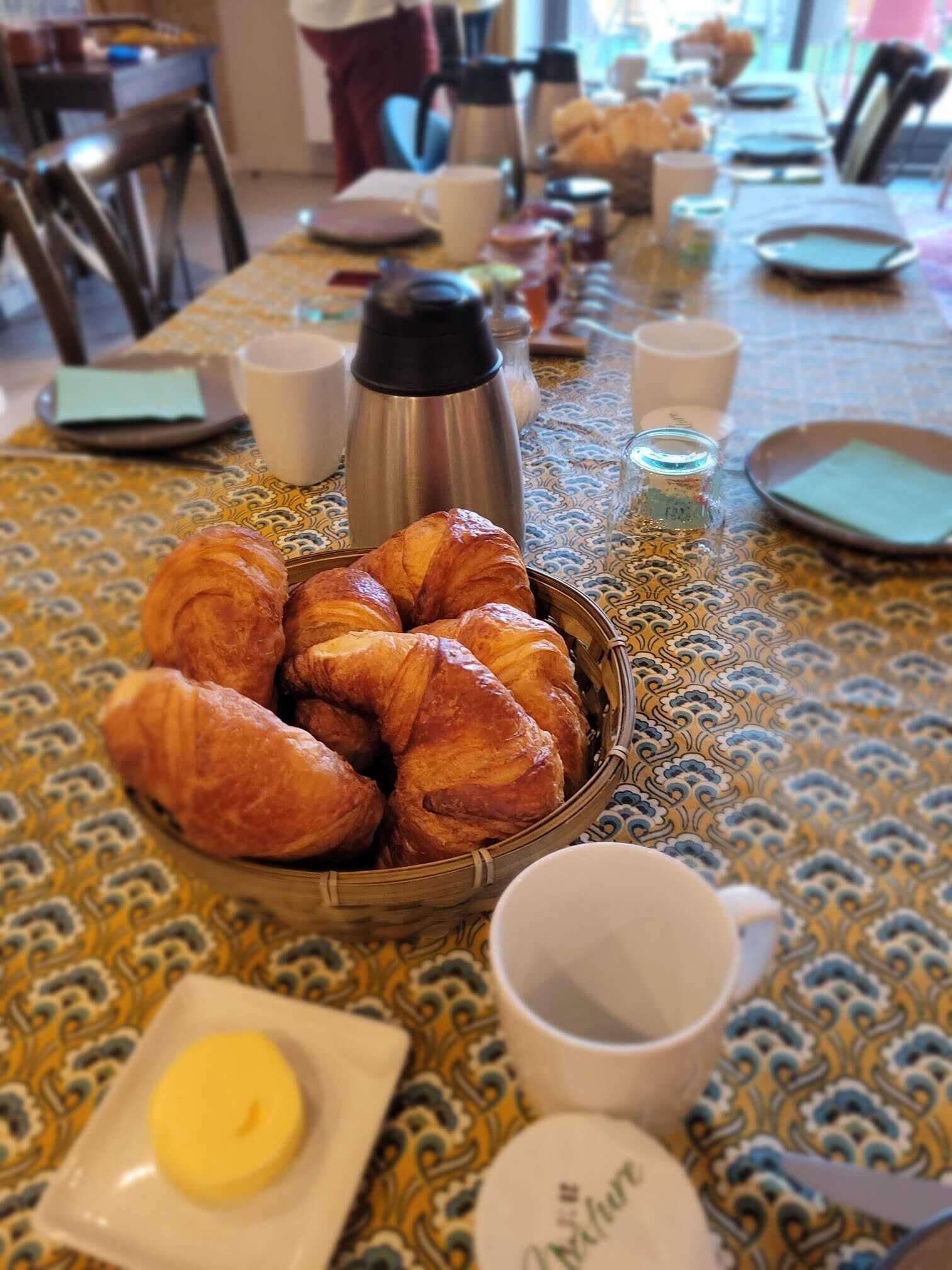 A table with a basket of croissants, a thermos, cups, and a plate with butter.