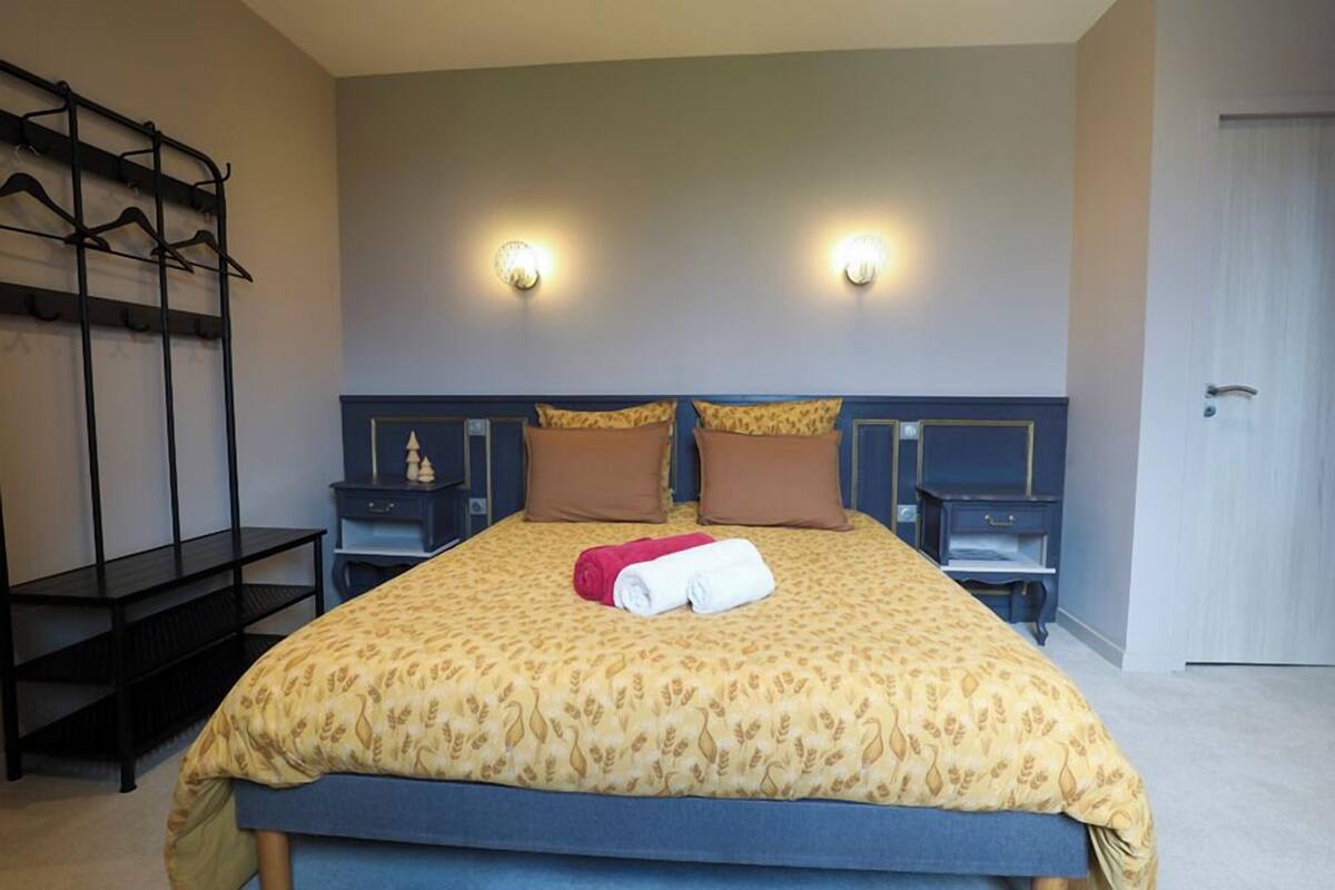 Un lit bien fait, recouvert d’une couvre-lit à motifs, deux tables de chevet avec des lampes, un porte-manteau et une porte, dans une chambre d’hôtel.