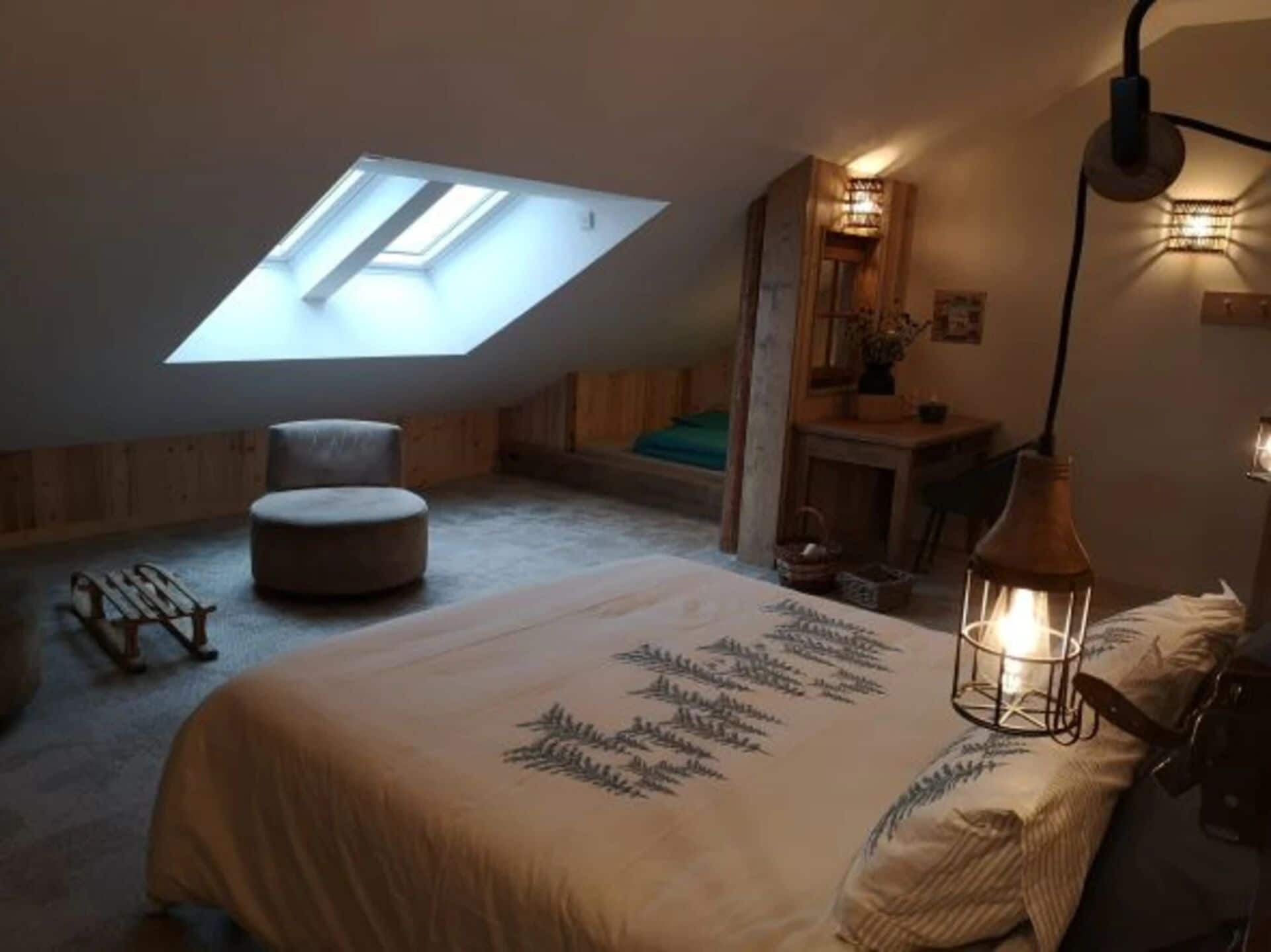 Een gezellig slaapkamer onder de dakpannen met dakraam, een rond voetenbankje, een bed met een bedovertrek met bladmotief en een hanglamp.