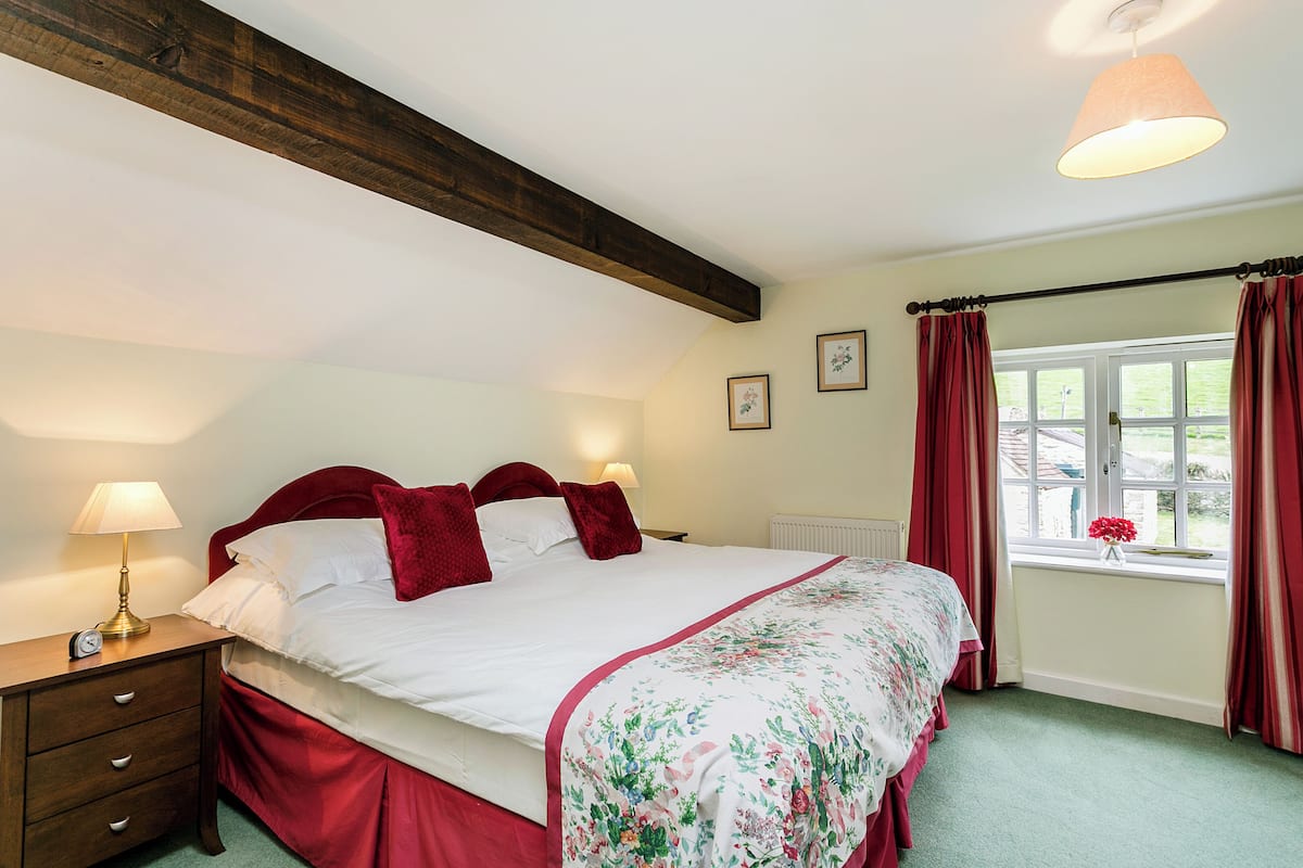 A neatly made bed with a floral bedspread, a wooden nightstand with a lamp, and a window with curtains.
