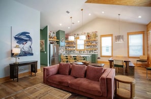 A spacious living room with a large sofa, a dining area with a table and chairs, a kitchen with a wooden island, and a ceiling with exposed beams.