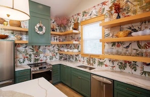 Una cocina con armarios verdes, estufa, lavaplatos y ventana con persianas.