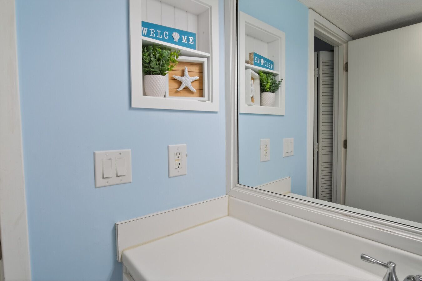 A bathroom with a white sink, a mirror, and a blue wall. There are two shelves with decorative items, including a starfish and a plant.