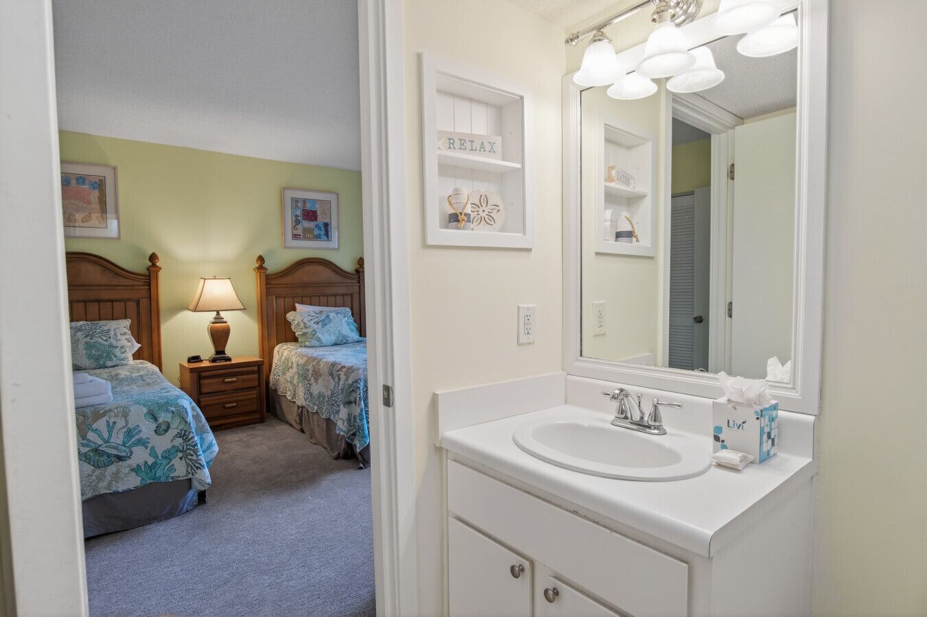 A bedroom with two beds, a nightstand with a lamp, a white bathroom with a sink and mirror, and a shelf with a 'RELAX' sign.