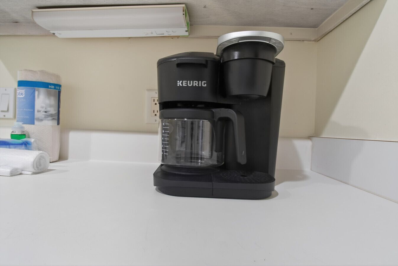 A black Keurig coffee maker on a white countertop in a room with a white wall and a ceiling light.