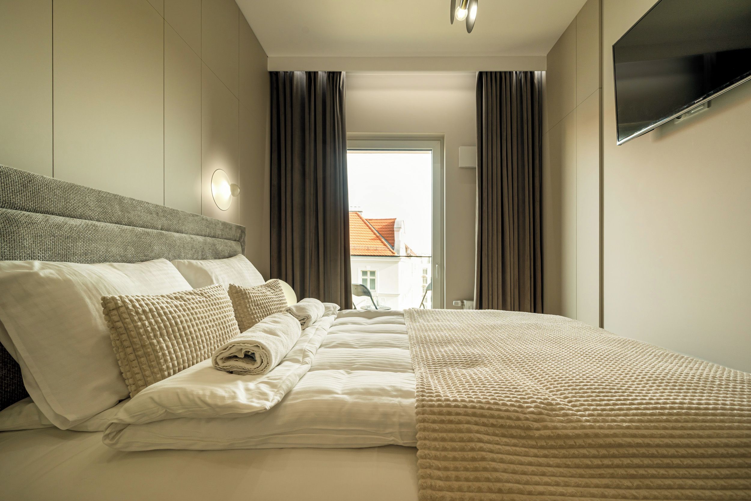 Habitación de hotel con cama, almohadas, una colcha, televisor de pantalla plana y ventana con cortinas.