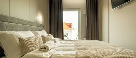 Habitación de hotel con cama, almohadas, una colcha, televisor de pantalla plana y ventana con cortinas.