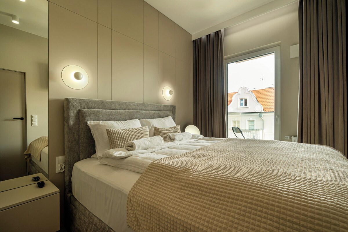 A modern hotel room with a large bed, a bedside table, and a view of the cityscape through the window.