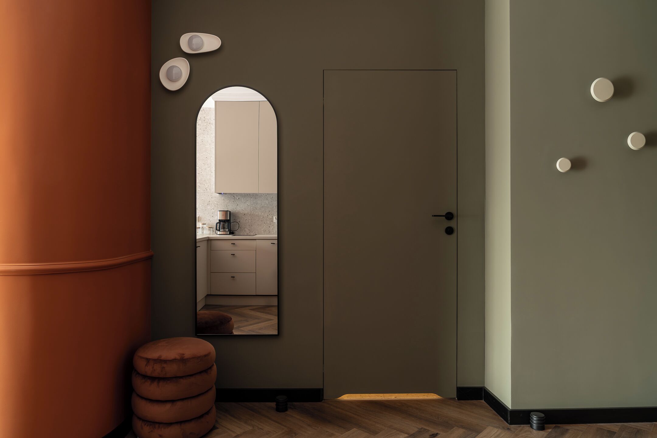 A modern interior with a wooden floor, a large cylindrical column, a door, and a mirror reflecting a kitchen area.