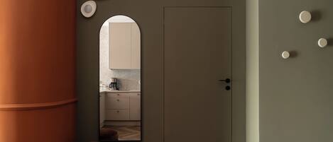 A modern interior with a wooden floor, a large cylindrical column, a door, and a mirror reflecting a kitchen area.