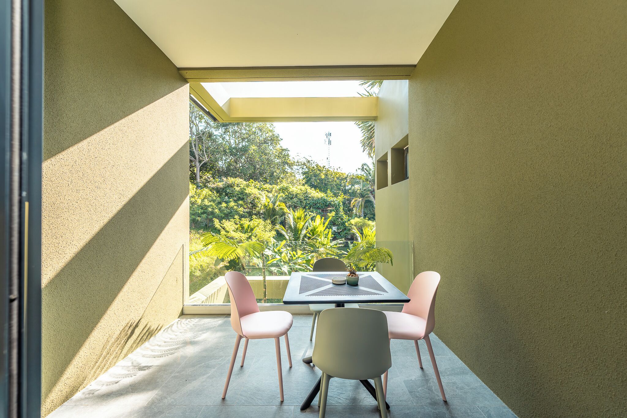 A modern balcony with a table and chairs, a potted plant, and a view of lush greenery.