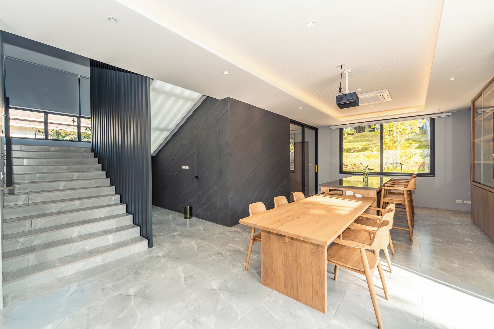 A modern interior with a staircase, a large wooden table, and a view of the outdoors.