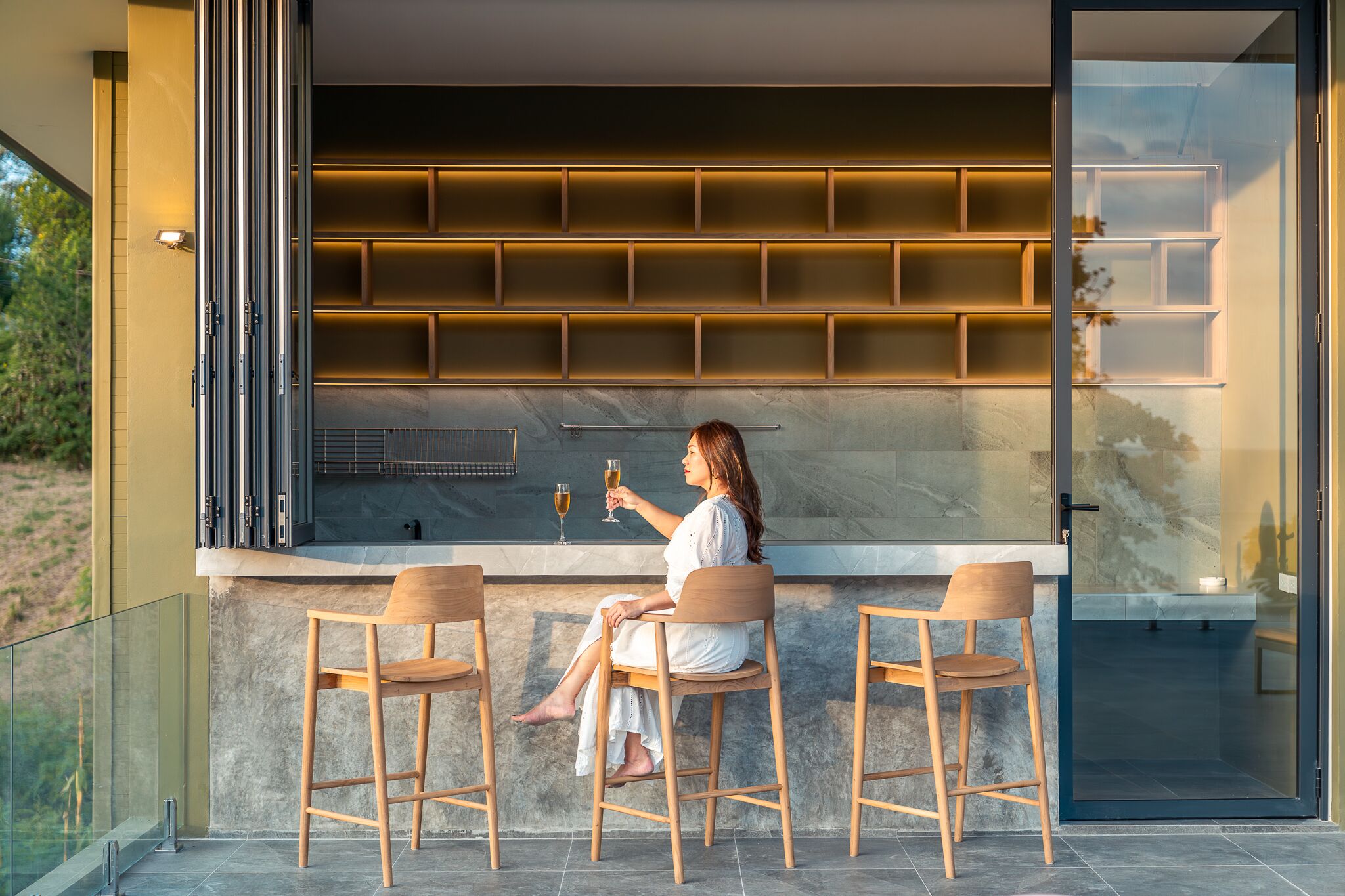 A woman sitting at a bar with two stools, holding a glass. The bar has shelves above and a glass railing to the left.