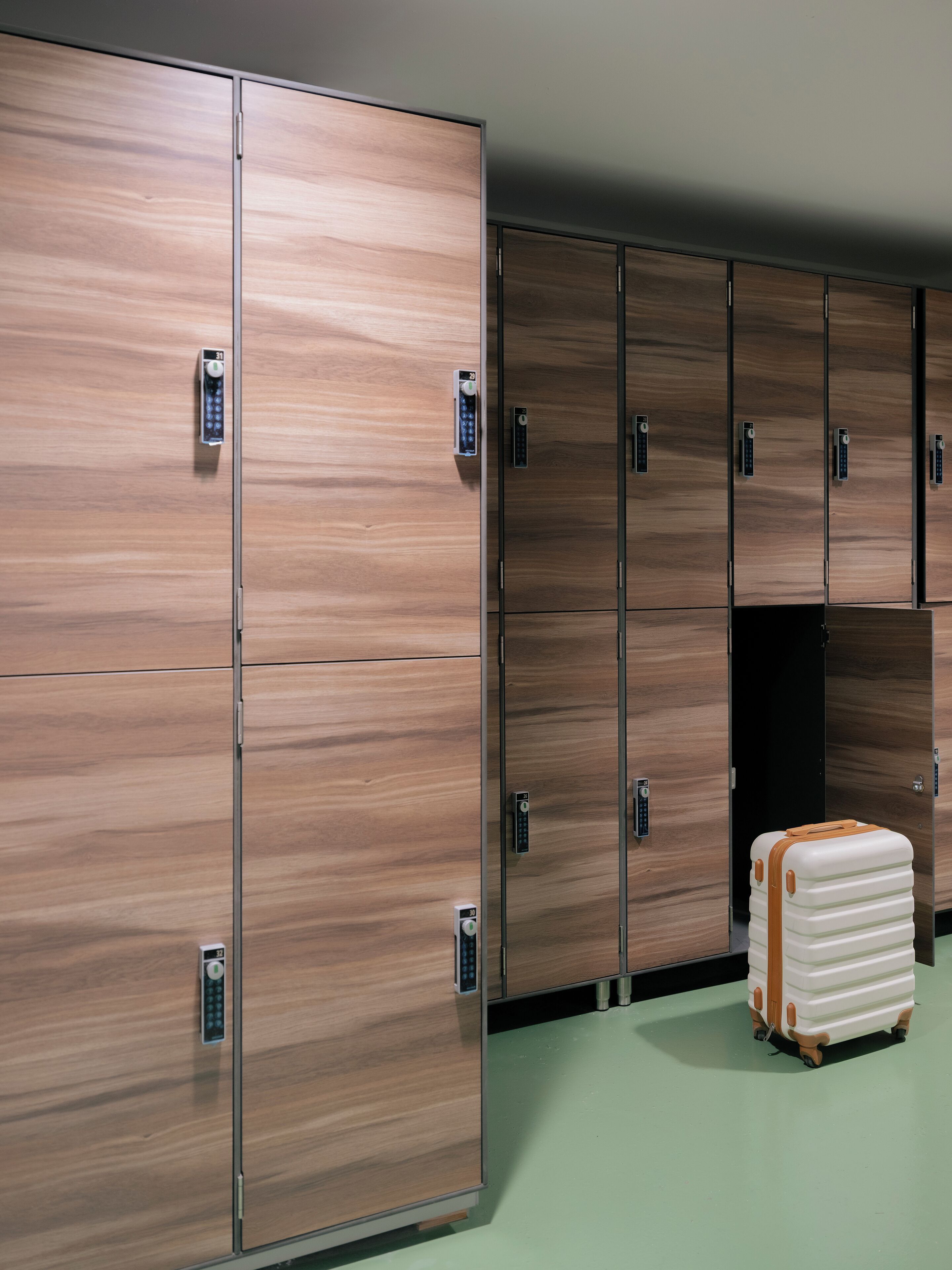 A row of wooden lockers with electronic keypads, a green floor, and a suitcase in the foreground.