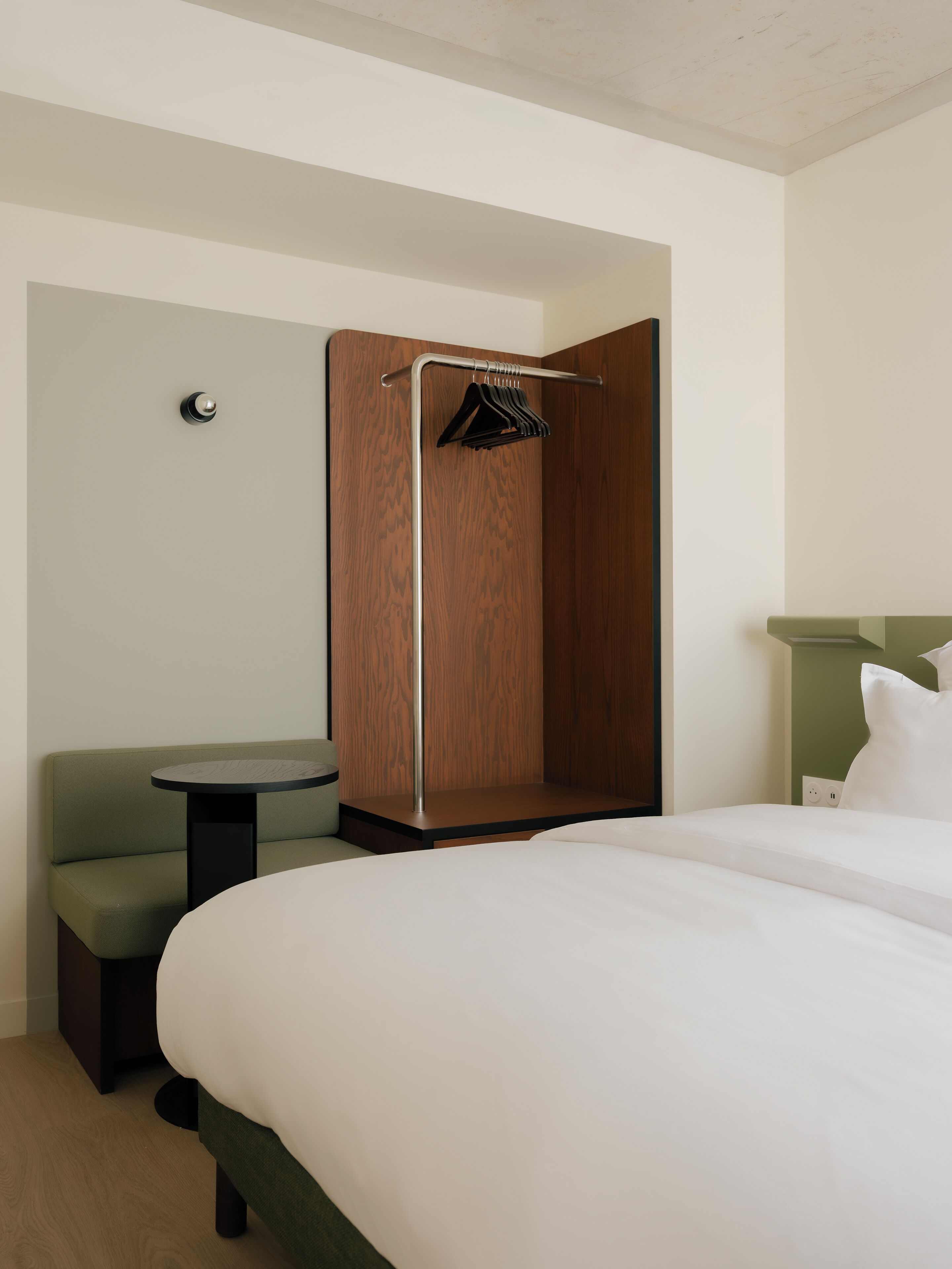 Une chambre d’hôtel comprenant un lit, une petite table, un banc et une armoire en bois avec une tringle pour suspendre les vêtements.