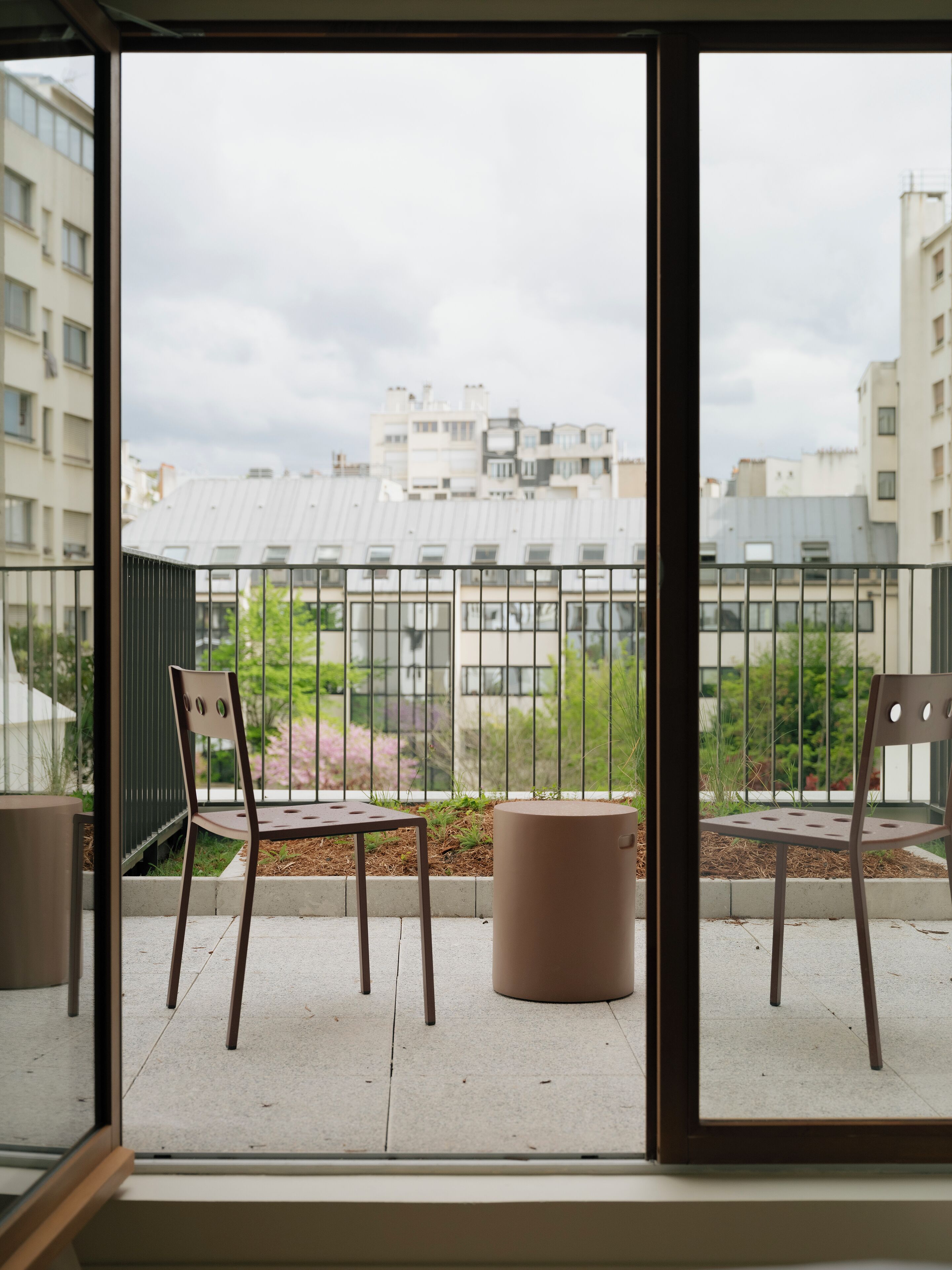En balkon med to stole og en skraldespand, der ud til et boligområde med bygninger og grønt.