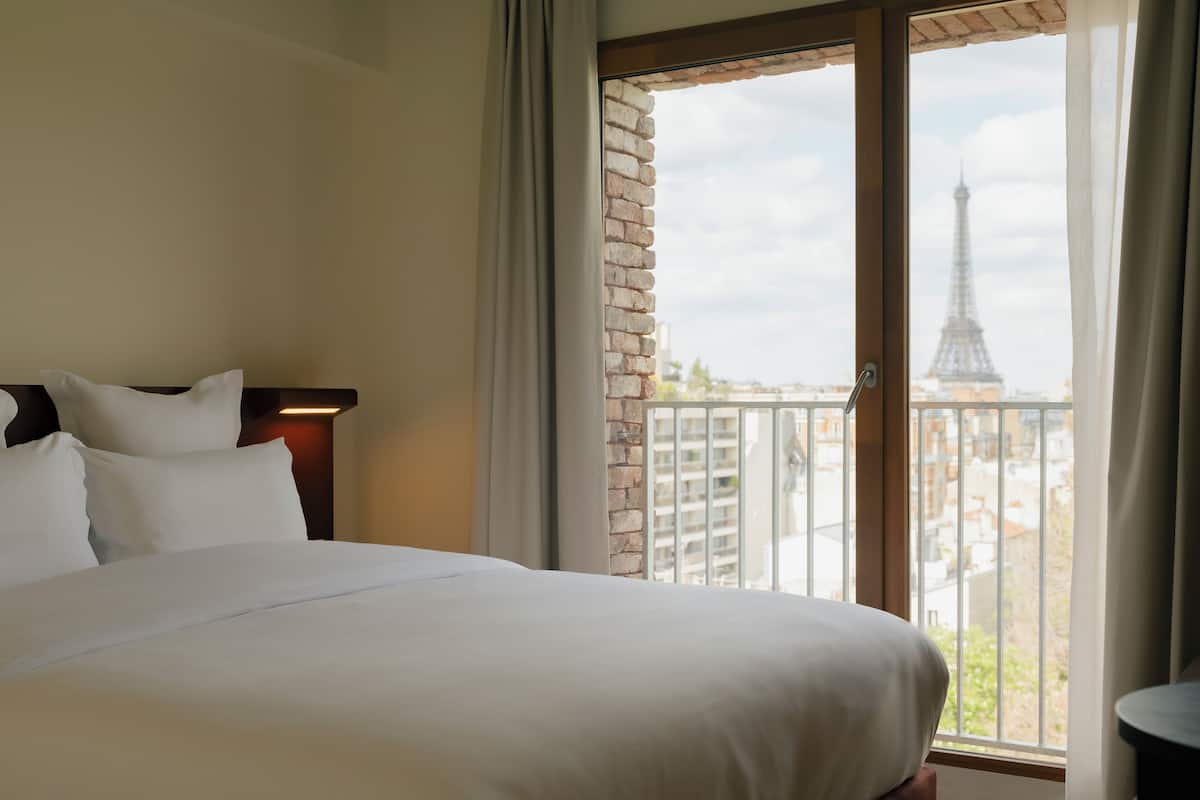 Quarto de hotel com uma cama grande, vista para a Torre Eiffel e uma varanda com guarda-corpo.
