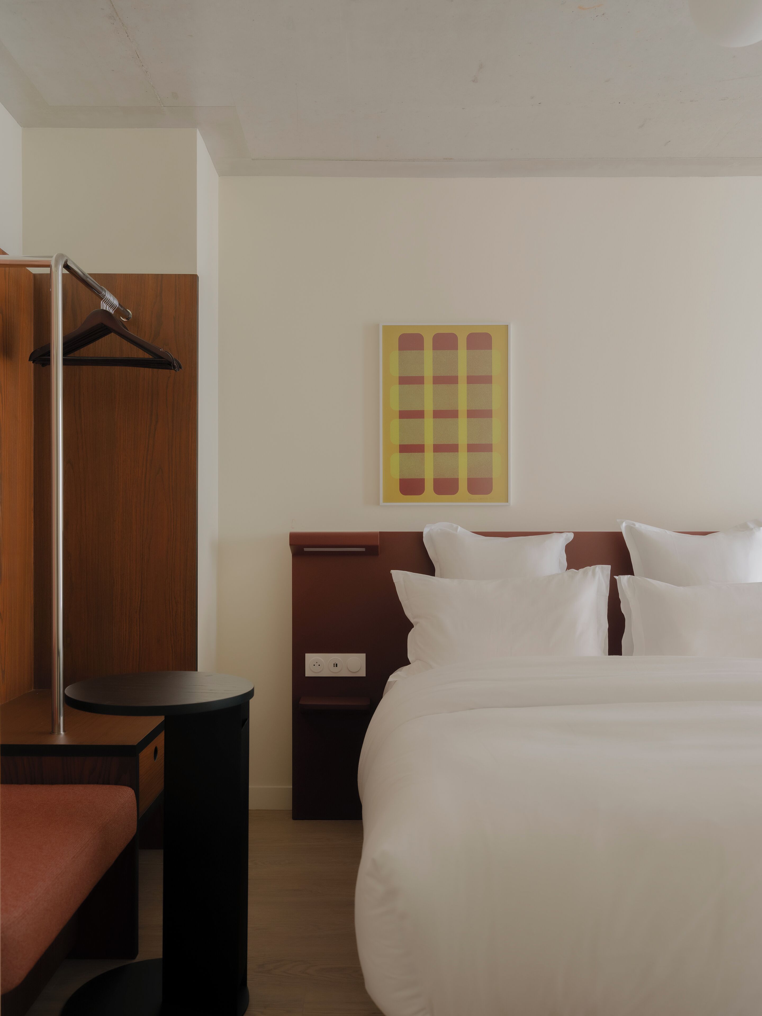 A hotel room with a large bed, a wooden wardrobe, a bedside table, and a wall-mounted artwork.