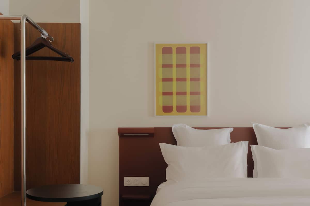 Quarto de hotel com uma cama grande, um guarda-roupa de madeira, uma mesa de cabeceira e uma obra de arte na parede.
