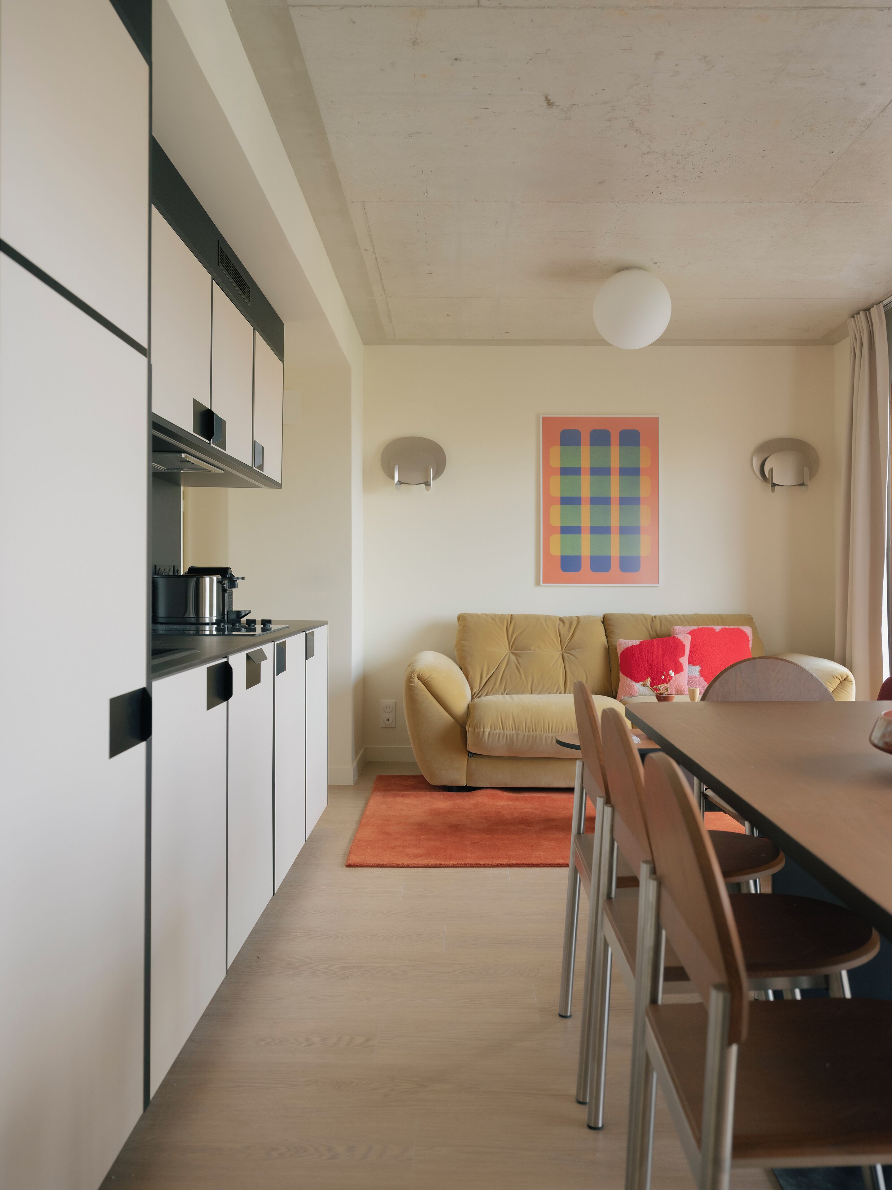 Een moderne keuken met witte kasten en een zwart aanrecht, een beige bank met een rode kussen, en een houten eettafel met stoelen.