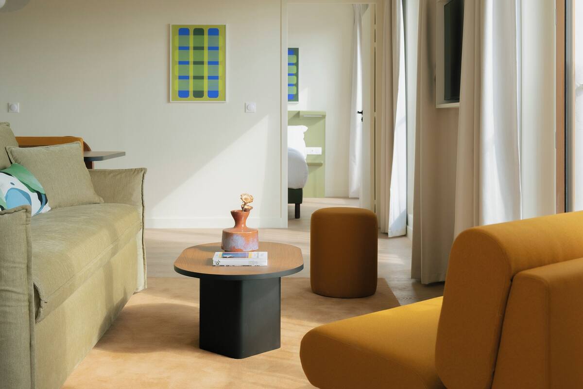Uma sala de estar moderna com um sofá, um banquinho e uma mesa de centro. Um corredor leva a um quarto.