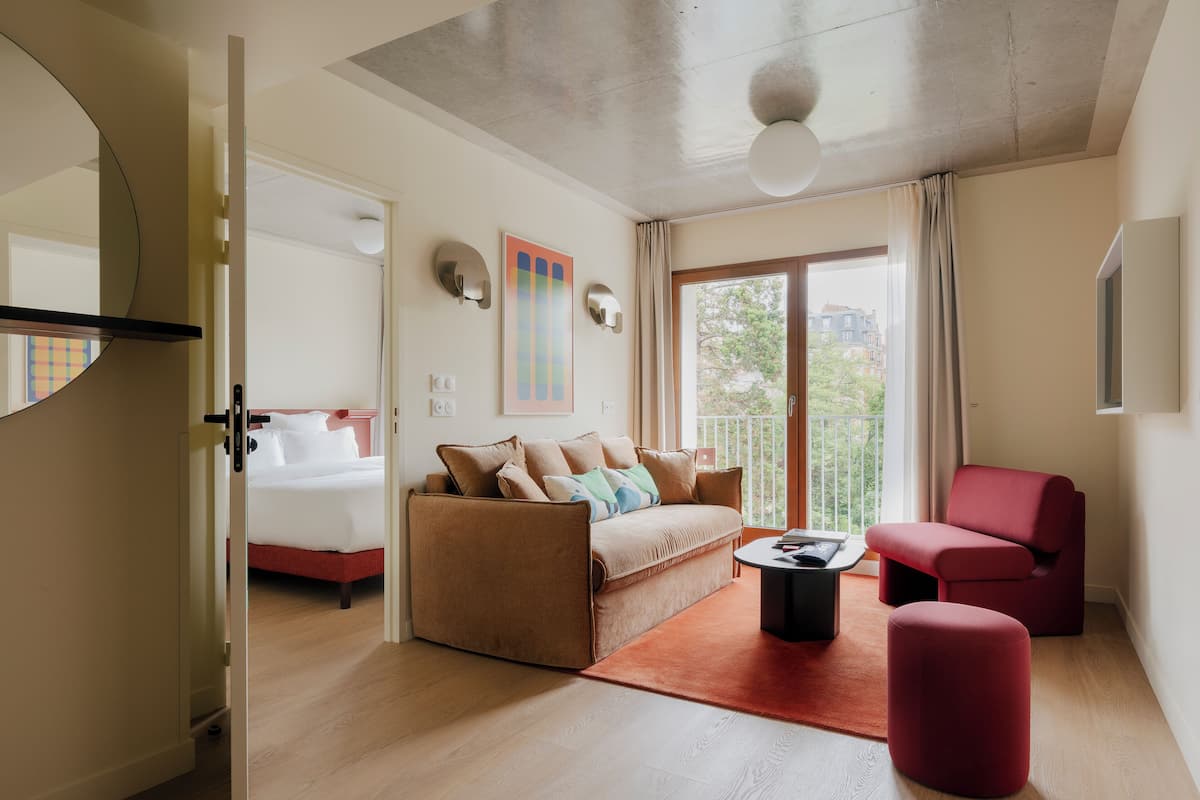 Quarto de hotel moderno com sofá, poltrona vermelha, mesinha e cama com cabeceira vermelha.