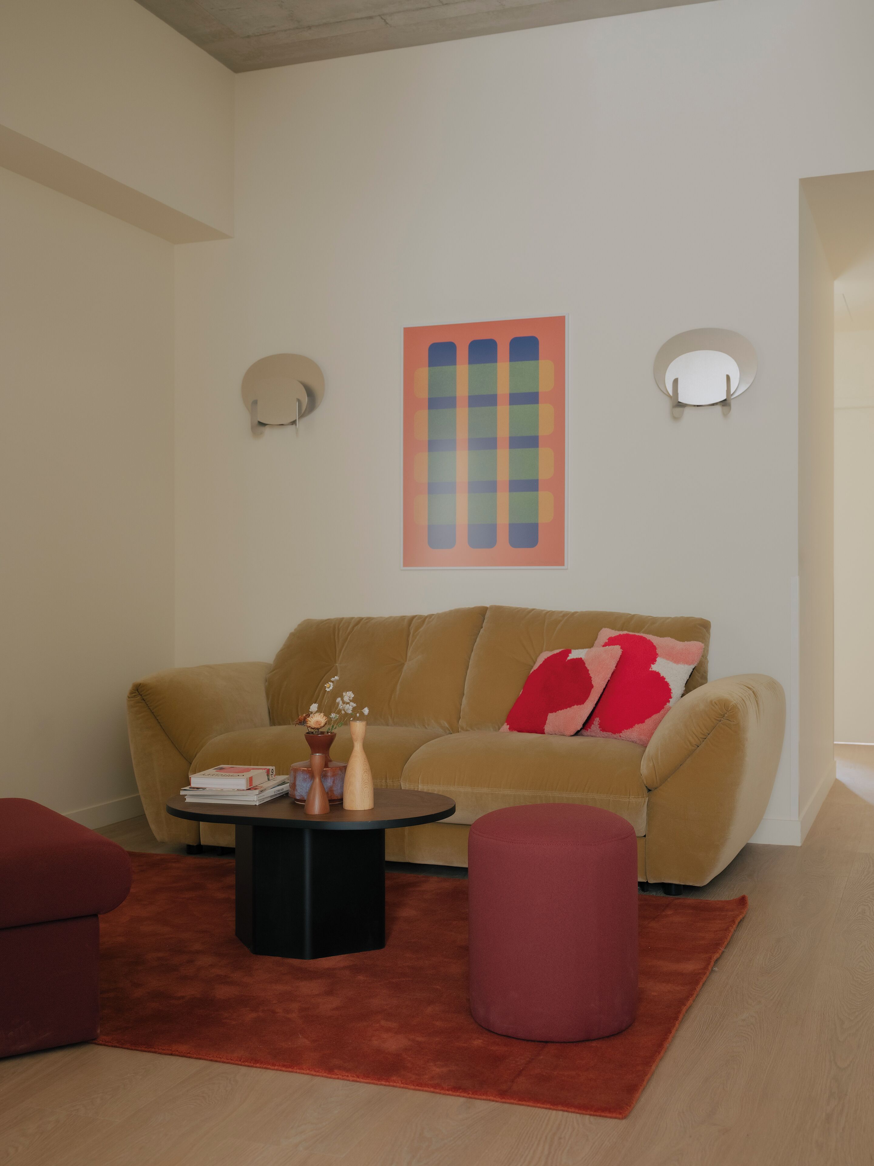 Een woonkamer met een beige bank, een ronde salontafel, een rode poef en een kleurrijk abstract schilderij aan de muur.