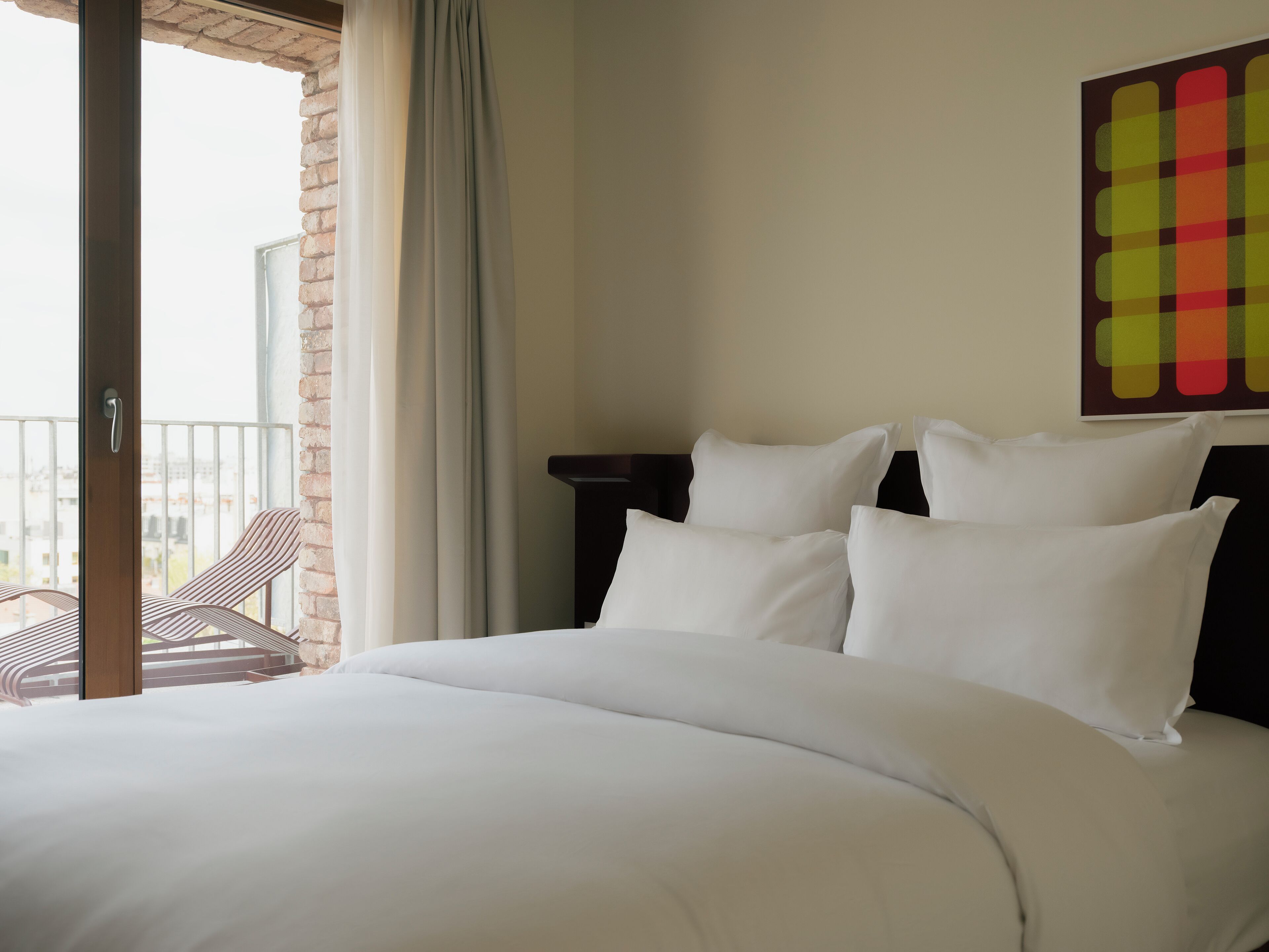 A hotel room with a bed, a view of the city, and a balcony with a chair.