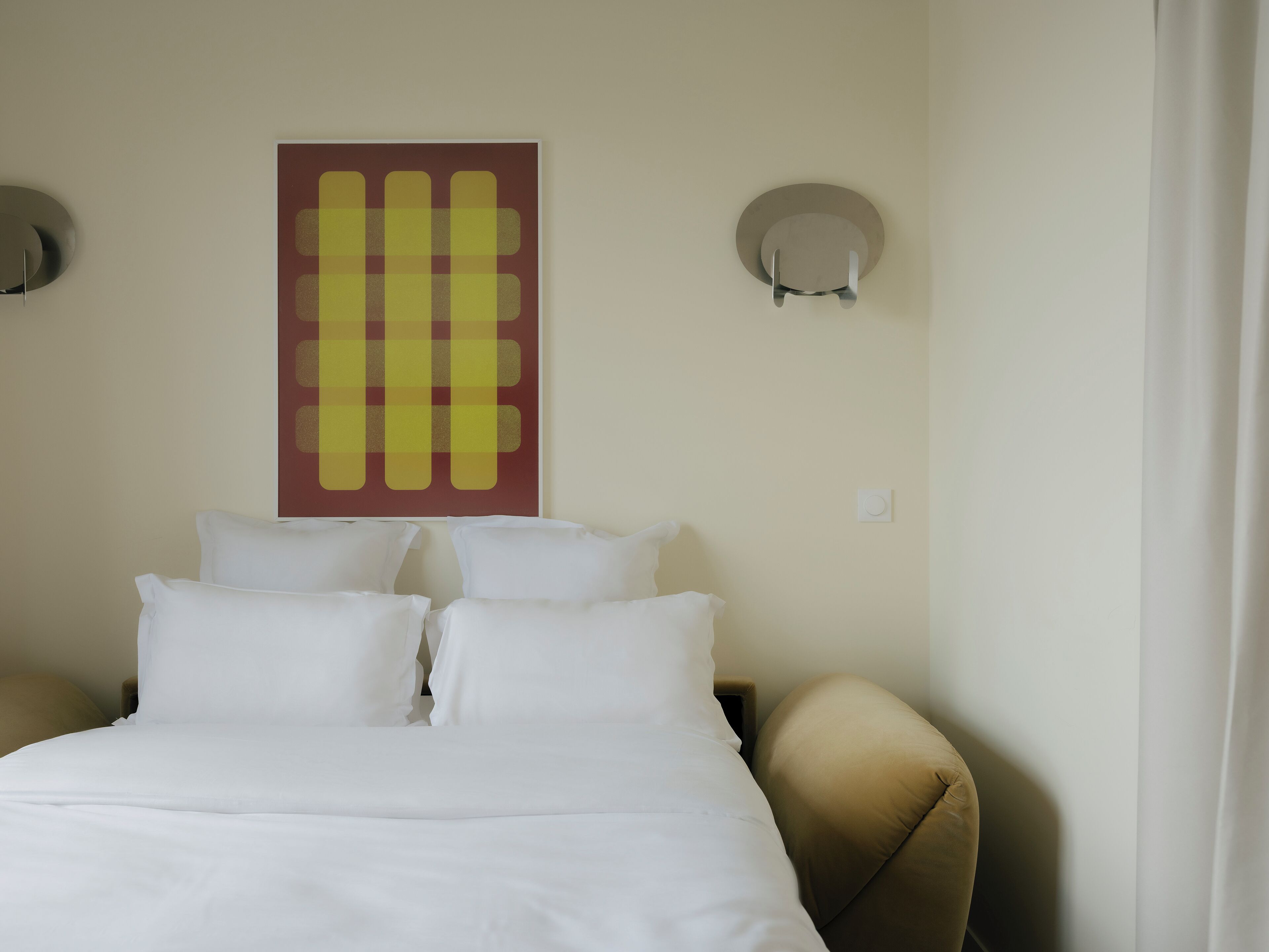 Une chambre d’hôtel avec un lit, des oreillers blancs, un fauteuil beige et une œuvre d’art accrochée au mur.