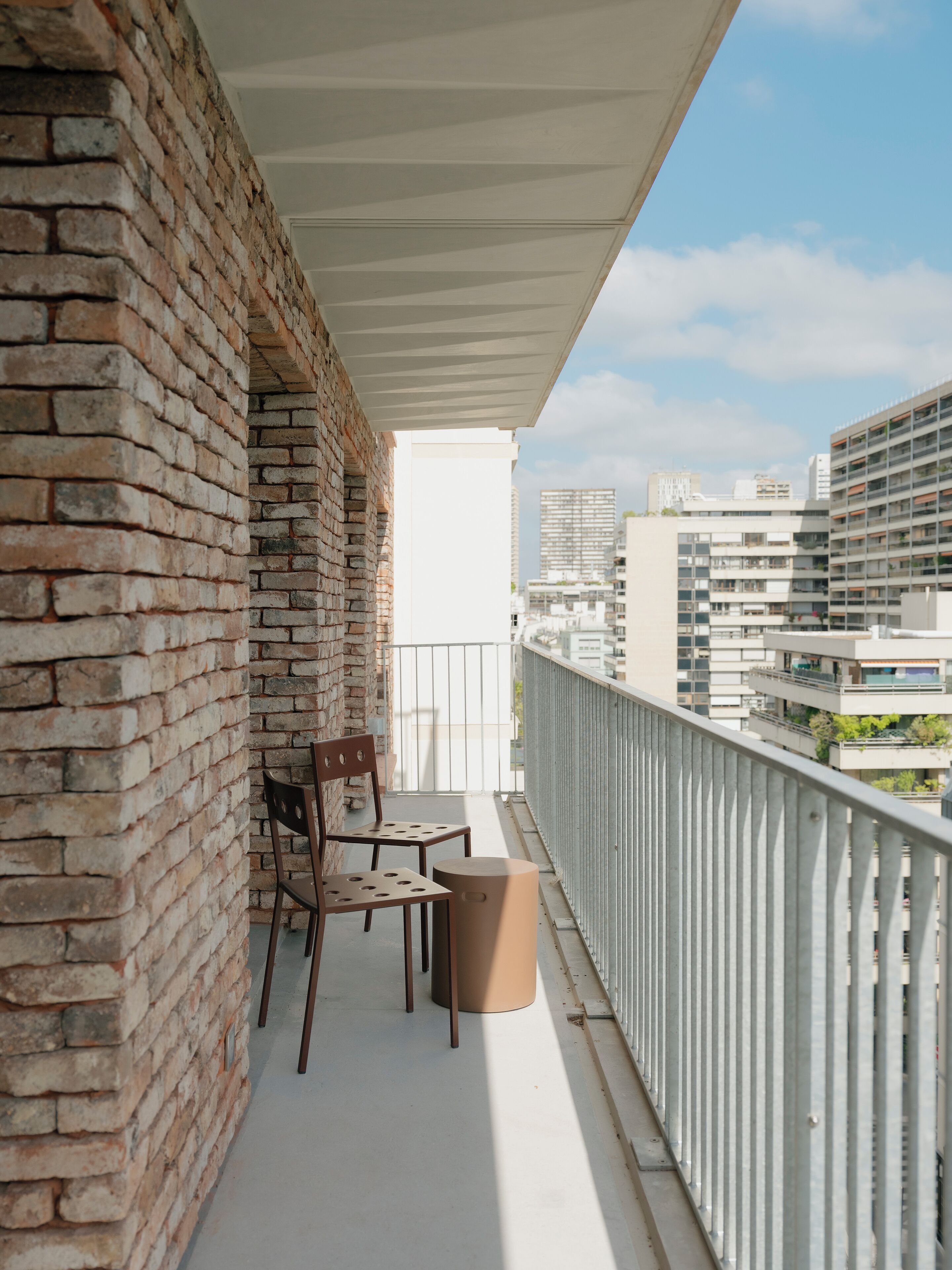 A balcony with a brick wall, two wooden chairs, a small round table, and a metal railing.