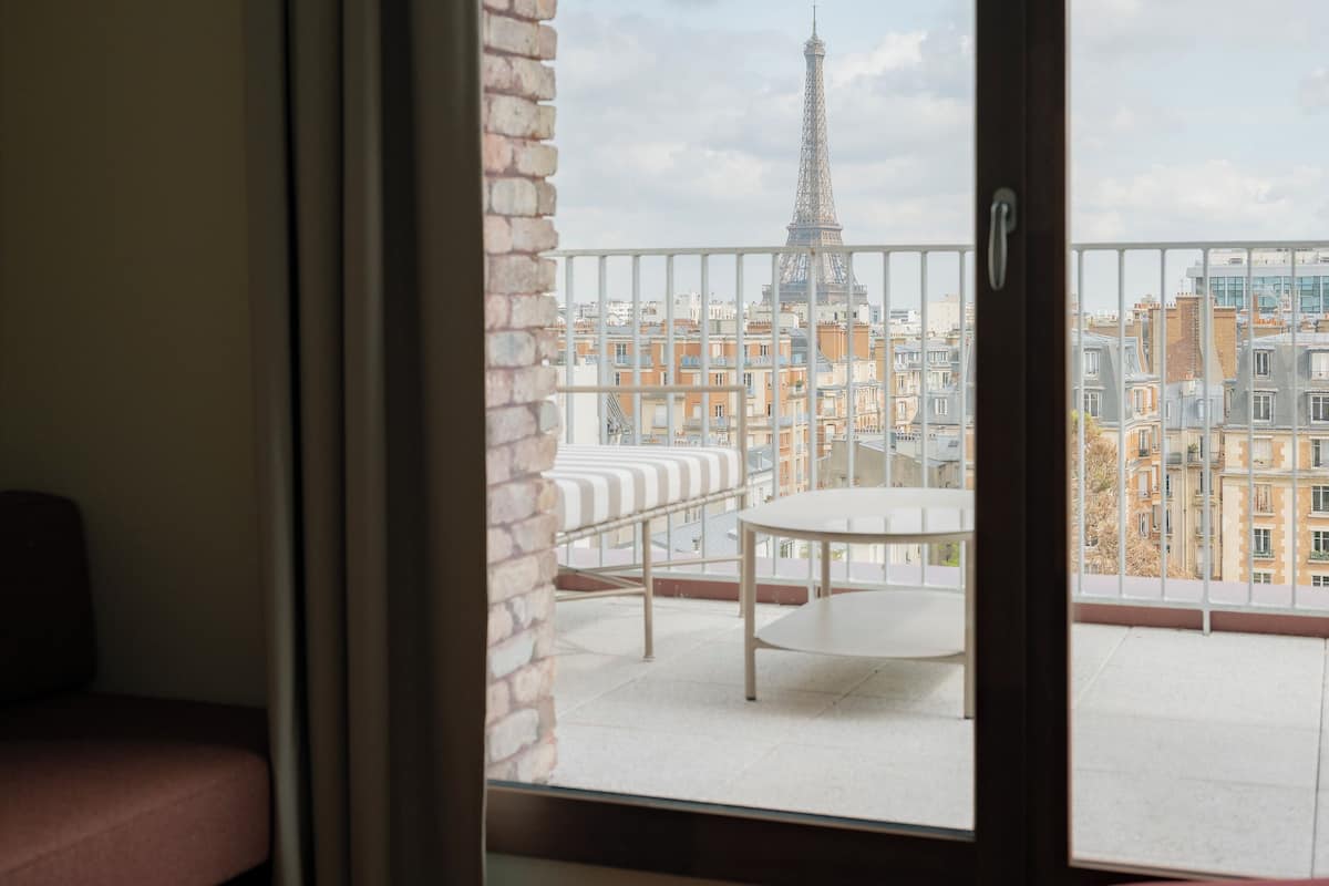 Quarto com vista para a Torre Eiffel através de uma porta de vidro, um banco branco e uma poltrona vermelha.