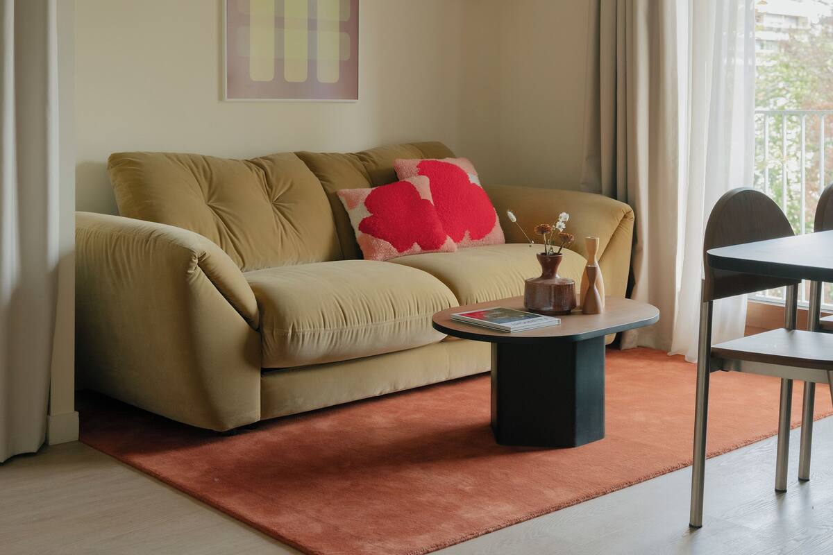 Uma sala de estar com um sofá bege, uma mesa de centro redonda e um tapete vermelho.