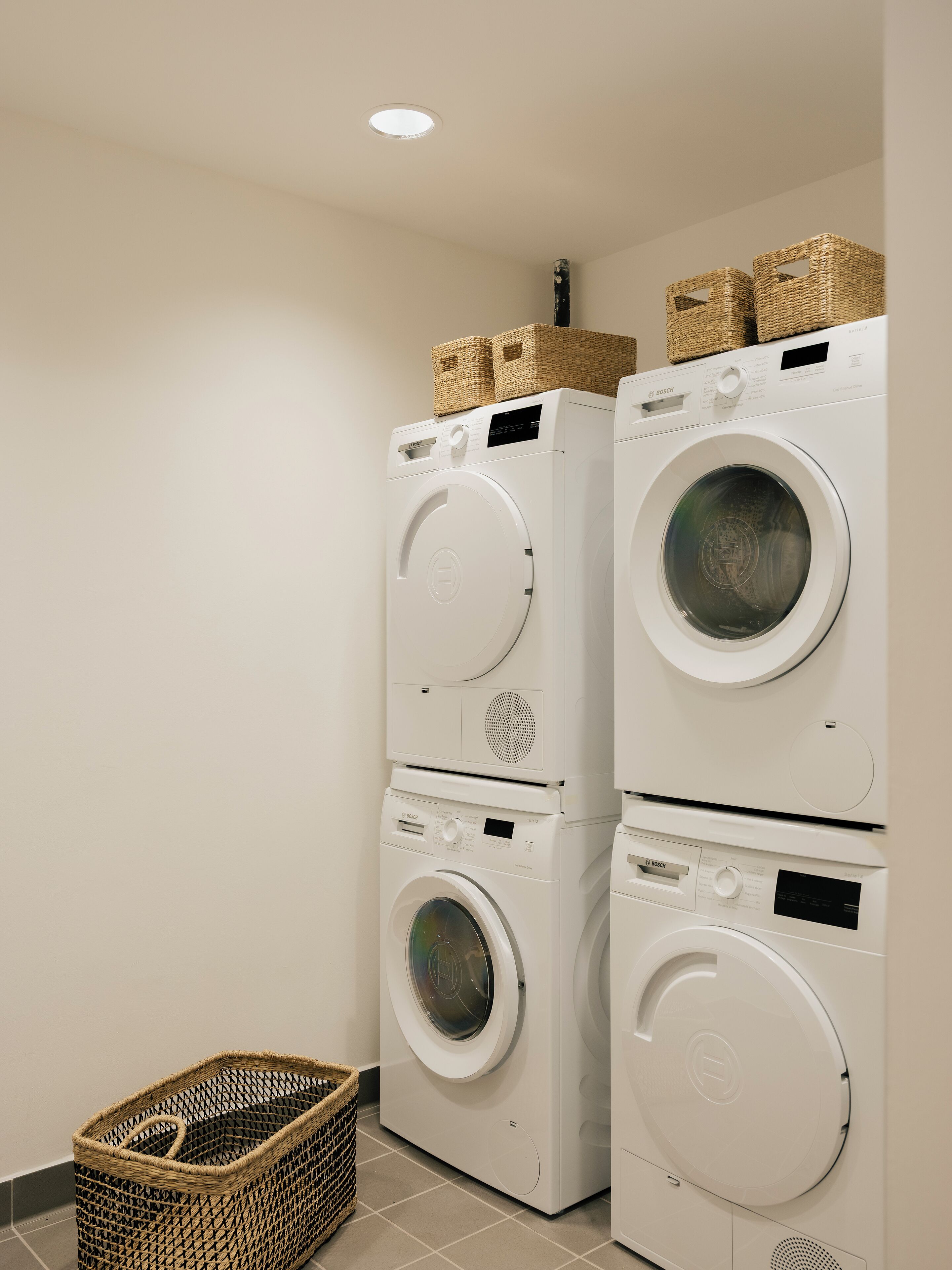 A laundry area with two stacked front-loading washing machines, a wicker basket, and a tiled floor.