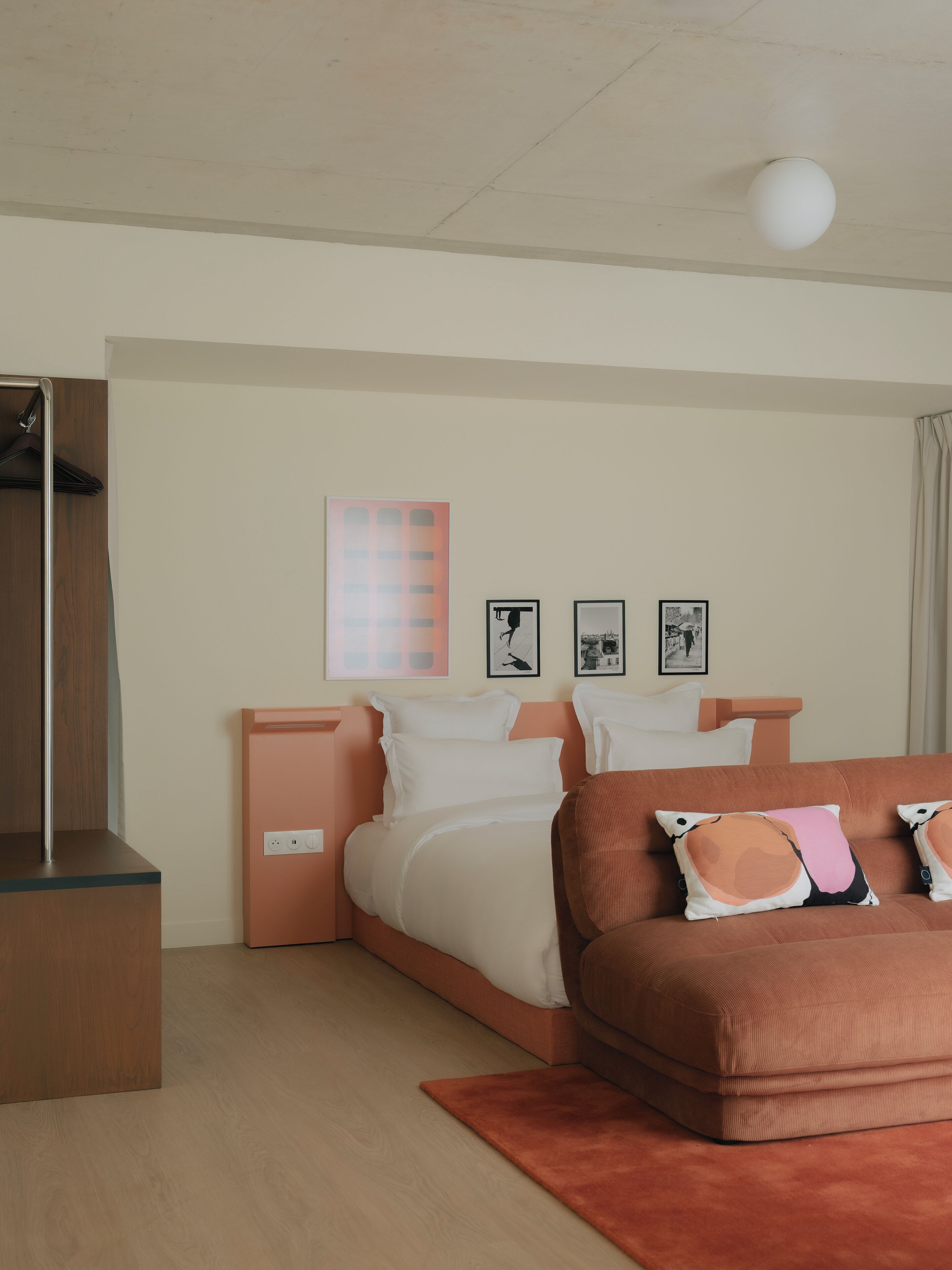 A modern bedroom with a wooden floor, a bed with white bedding, a brown sofa with patterned cushions, and a ceiling light.