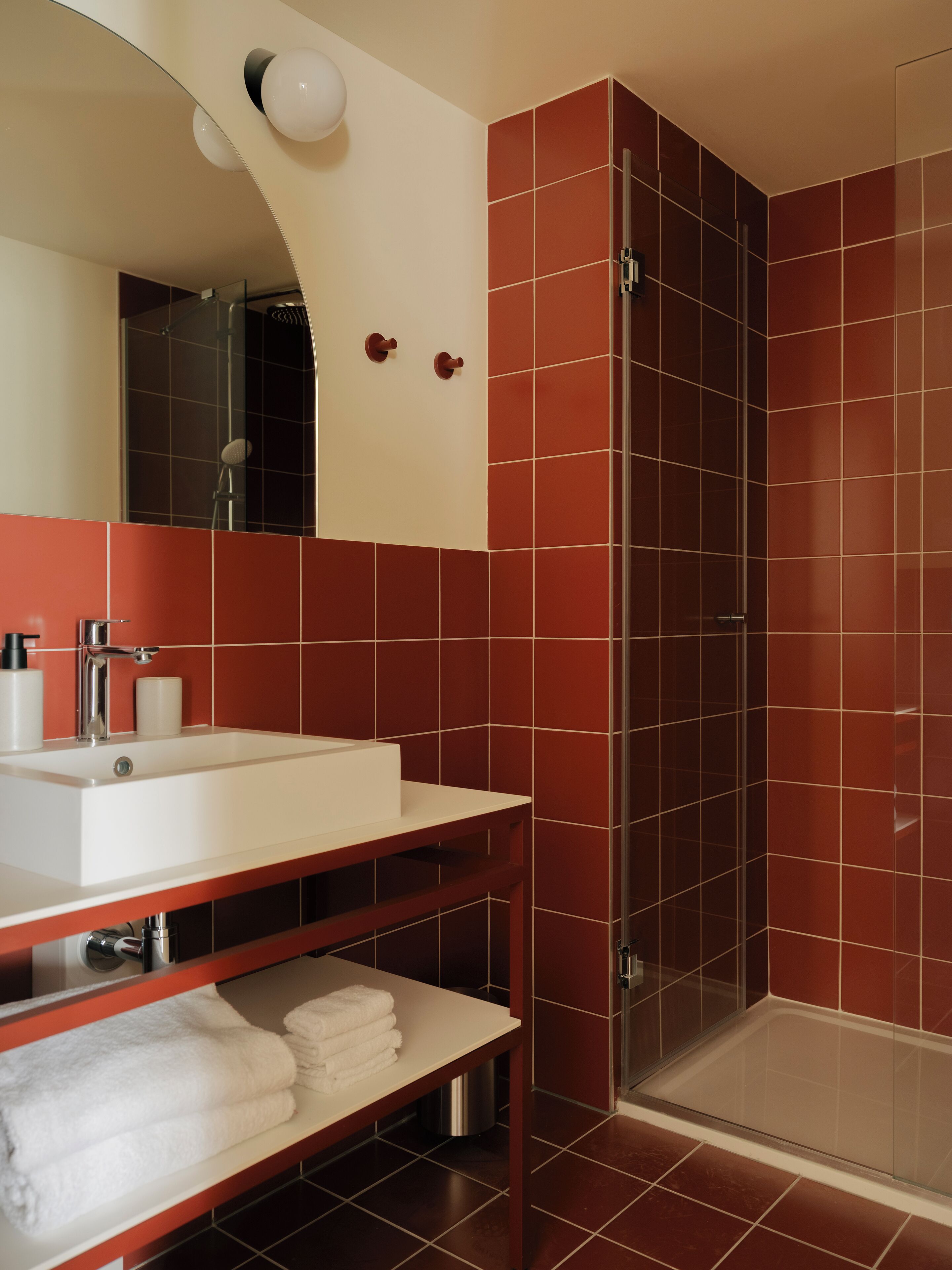 Une salle de bain moderne avec un lavabo blanc, des murs recouverts de carreaux rouges, une douche en verre et un miroir au-dessus du lavabo.