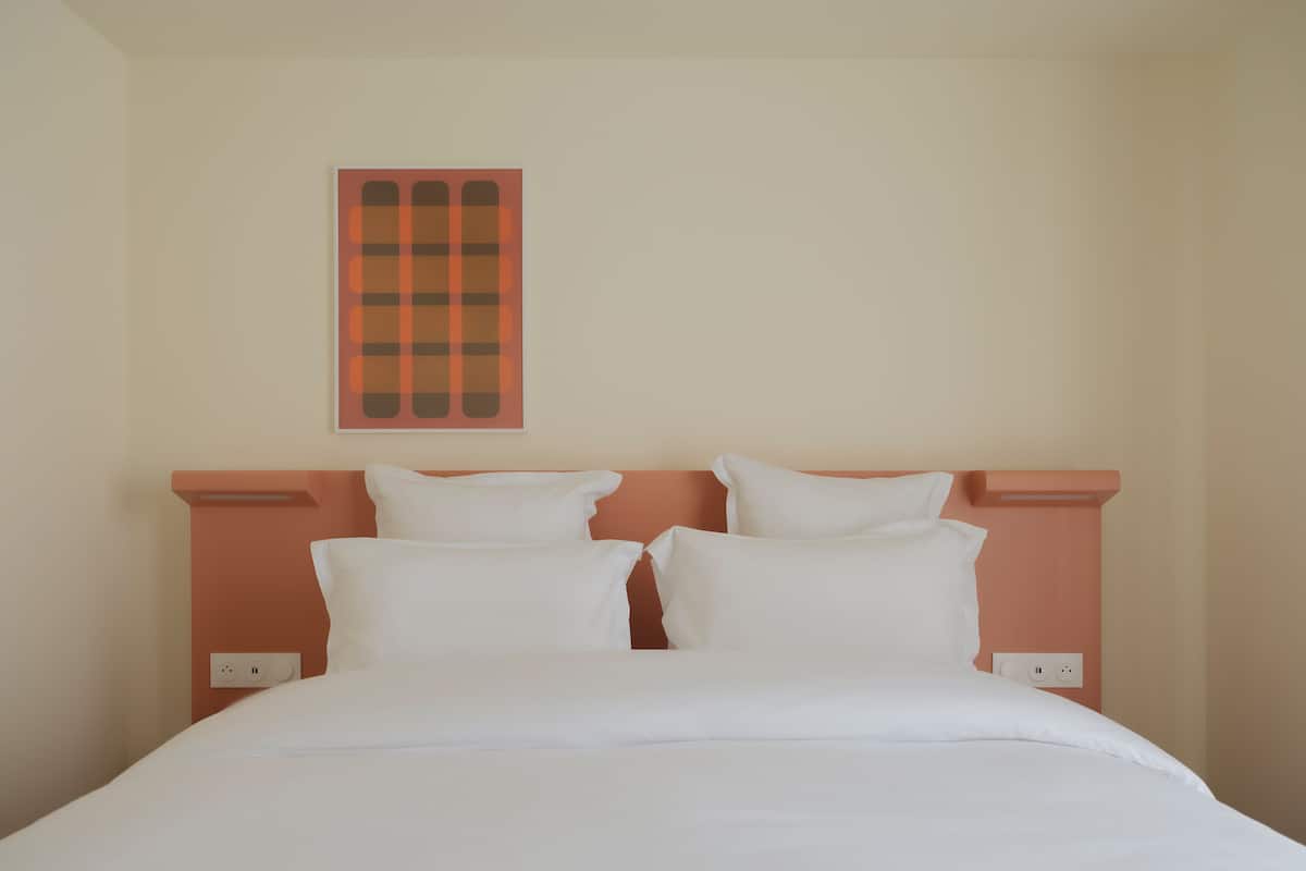 Uma cama com roupa de cama branca, cabeceira rosa e uma obra de arte pendurada na parede.