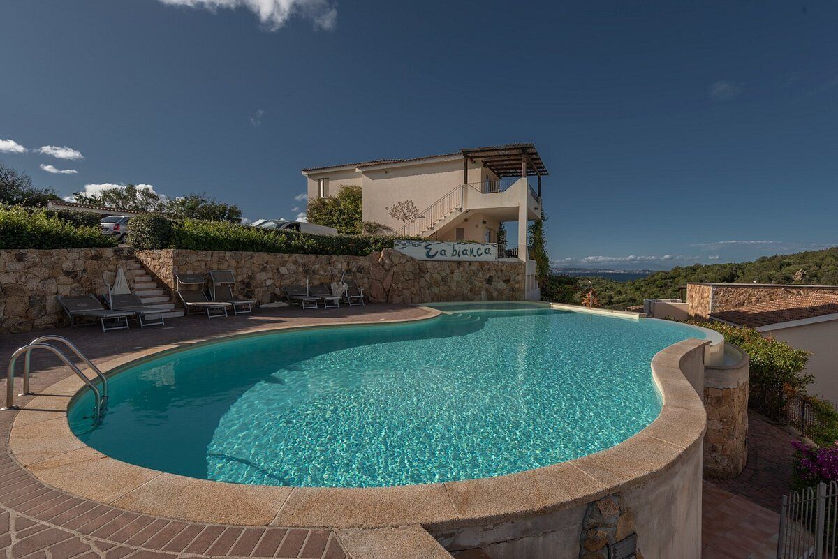 A swimming pool with a stone wall and lounge chairs, a building with a sign reading "La Bianca", and a clear blue sky.
