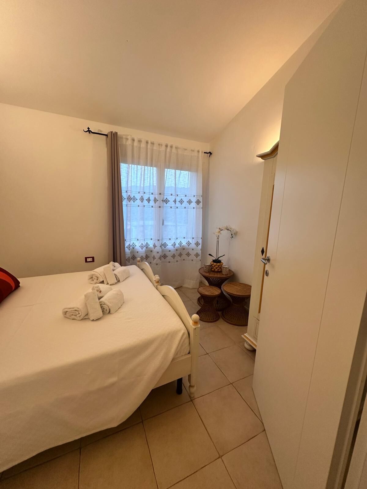A neatly made bed with white linens and rolled towels, a small round table with a vase of flowers, and a door to the right.